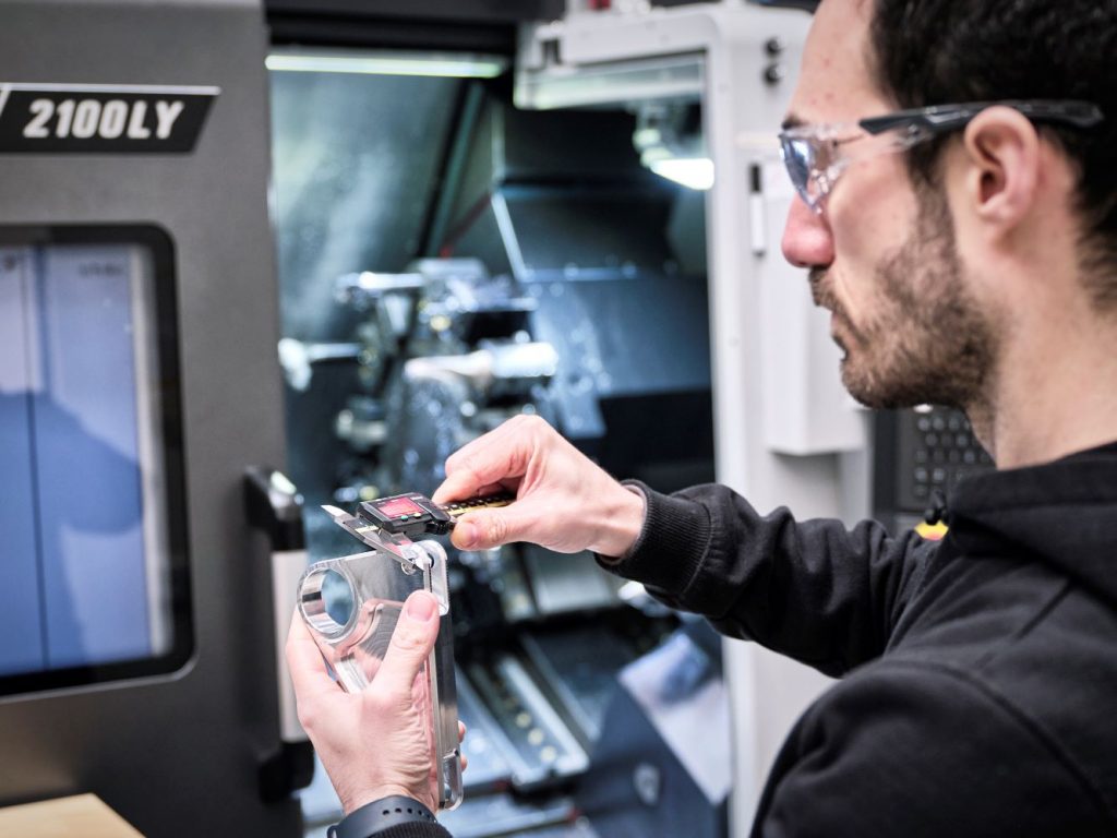 A man wearing safety glasses closely inspecting a metal component with a caliper, with a CNC machine labeled '2100LY' in the background.