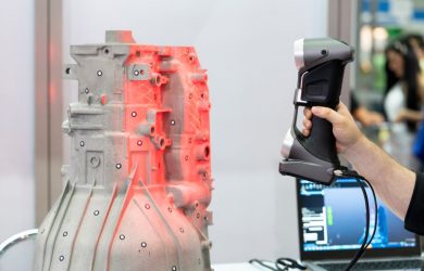 A hand holding a 3D scanner aimed at a complex metal object highlighted with red scanning lines.